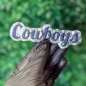 Puede incluir: Un logotipo de los Cowboys en plata brillante y azul marino. La palabra "Cowboys" está bordada en hilo azul marino sobre un fondo plateado brillante. El artículo se sostiene contra un fondo verde borroso.