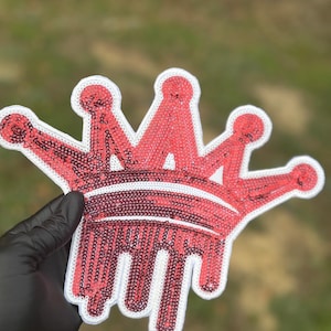 May include: A crown-shaped patch covered in red sequins with a white border. The design features a classic crown silhouette with five points, and the base of the crown has a dripping effect, with red sequined drips extending downward from the band.