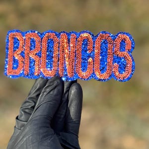 May include: A blue and orange glittered Broncos sign. The word "BRONCOS" is spelled out in orange glitter against a blue glitter background. The sign is held by a gloved hand.
