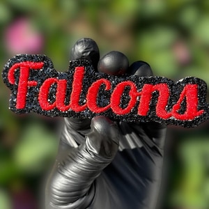 Könnte beinhalten: Ein schwarzes, glitzerndes Emblem mit dem Wort "Falcons" in Rot, vor einem verschwommenen grünen und rosa Hintergrund. Der Text ist in einer fetten, stilisierten Schriftart gehalten, mit einem glitzernden schwarzen Rand. Das Emblem ist wahrscheinlich ein Teamlogo oder eine Dekoration.