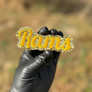 May include: A yellow embroidered "Rams" patch with a gold glitter border. The patch is held by a gloved hand against a blurred outdoor background. The lettering is in a cursive style.