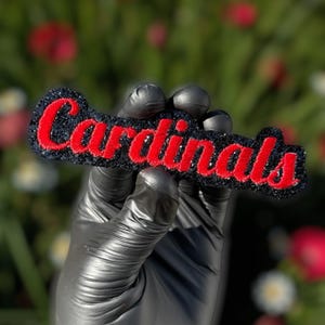 May include: A black and red Cardinals logo patch. The word "Cardinals" is embroidered in red on a black background. The patch is held in a black gloved hand against a blurred green and red floral background.