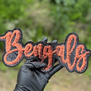 Puede incluir: Un parche decorativo que muestra la palabra Bengals en letras cursivas de lentejuelas naranjas. El texto está delineado en negro sobre un fondo de lentejuelas negras, creando un contraste marcado. El parche tiene un borde negro grueso.