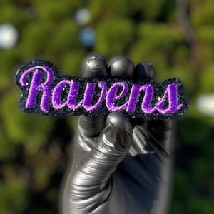 May include: A black patch with the word "Ravens" embroidered in purple. The patch has a black glitter border and is held by a gloved hand. The background is a blurred green.