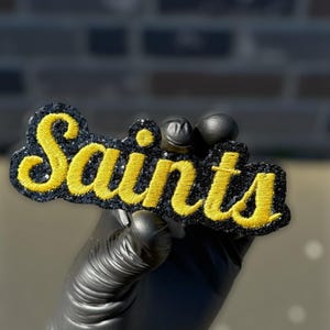 May include: A black and gold embroidered patch with the word "Saints" in yellow script. The patch has a glittery black border and is held up by a gloved hand. The background is a blurred brick wall.