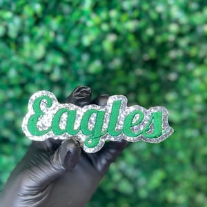May include: A decorative patch featuring the word Eagles in green embroidered script lettering. The text is outlined with a thick border of silver glitter material, creating a textured and sparkling appearance against the green background.