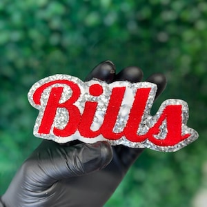 May include: A Bills-themed decorative item with a silver glitter border and backing. The word "Bills" is written in bold red letters. The item is held by a hand wearing a black glove against a blurred green background.