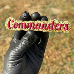 Puede incluir: Una barra dorada con purpurina con la palabra "Commanders" en letras burdeos. La barra está sujeta por una mano enguantada sobre un fondo exterior borroso. El artículo es probablemente un accesorio decorativo o un emblema.