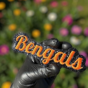May include: A black and silver glitter patch with the word "Bengals" embroidered in orange. The patch is held by a gloved hand against a blurred floral background. The patch is a sports-themed accessory.