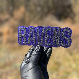 May include: A purple and black glittery Ravens emblem. The word "RAVENS" is spelled out in a stylized font, with a black outline. The emblem is held up against a blurred outdoor background.