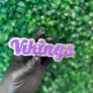 May include: A decorative patch featuring the word Vikings in purple embroidered script lettering. The text is outlined by a thick, textured border covered in silver glitter, creating a high-contrast appearance against the purple fabric letters.