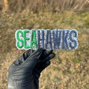 May include: A beaded barrette with the word "SEAHAWKS" in green, blue, and silver. The barrette is held by a gloved hand against a blurred outdoor background. The letters are outlined with black beads.