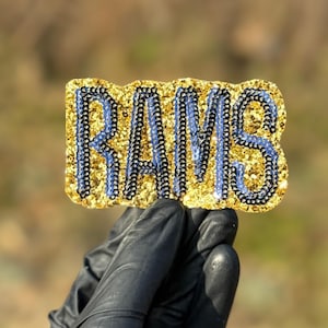 Könnte beinhalten: Ein rechteckiges, glitzerndes goldenes Emblem mit dem Wort "RAMS" in Blau, umrandet von Schwarz. Das Emblem wird von einer behandschuhten Hand vor einem verschwommenen Hintergrund im Freien gehalten. Das Design ist wahrscheinlich für ein Sportteam.