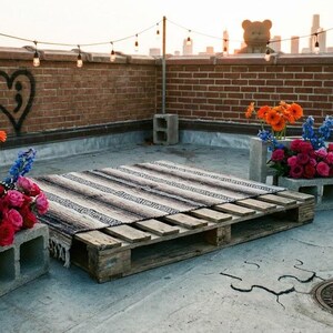 May include: A rooftop scene with a wooden pallet, a striped blanket, and concrete block planters filled with colorful flowers. A brick wall with a heart and semicolon graphic is in the background, along with string lights and a teddy bear.