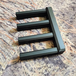 May include: A black, geometric object with a rectangular frame and horizontal bars. The object is made of a matte material and sits on a camouflage-patterned surface. The design is modern and minimalist.
