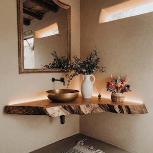 Puede incluir: Una esquina de baño con una estantería de madera de borde vivo, un lavabo dorado en forma de cuenco y un jarrón blanco con flores azules. Un espejo decorativo cuelga sobre el lavabo. La estantería tiene una tira de luz cálida.