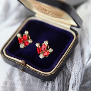 Peut inclure: Une paire de boucles d'oreilles vintage avec des pierres précieuses rouges et claires, présentée dans un écrin noir avec un intérieur en velours bleu. Les boucles d'oreilles présentent un motif en grappe avec des pierres rectangulaires et rondes.