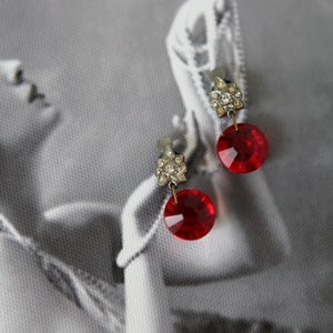 Może przedstawiać: Para zabytkowych kolczyków na klips. Każdy kolczyk ma mały, srebrny kwiatowy wzór z przezroczystymi dżetami, połączony z dużym, fasetowanym, okrągłym, rubinowo-czerwonym szklanym kamieniem. Tło to czarno-białe zdjęcie.