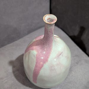 May include: A handcrafted ceramic vase with a rounded body and a slender neck. The vase features a light green base color with a prominent pink stripe design. The rim is a light brown color.