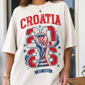 May include: An off-white short-sleeve t-shirt featuring a distressed red and blue graphic. The text reads CROATIA above the large numbers 2026, which frame a central trophy illustration containing a checkered pattern. Below is a banner with EST. 2026.