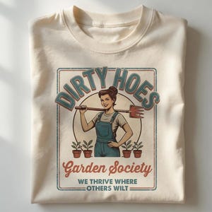 May include: A cream-coloured t-shirt with a vintage-style graphic. The design features a woman holding a gardening tool, the text "Dirty Hoes Garden Society", and the slogan "We thrive where others wilt".