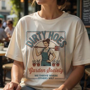May include: Cream-colored t-shirt featuring a vintage-style graphic. The design includes the words "DIRTY HOES" in a circular frame, with a woman holding a gardening tool. Below, it reads "Garden Society" and "WE THRIVE WHERE OTHERS WILT".