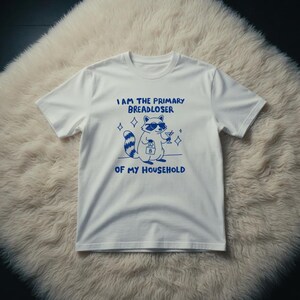 May include: White t-shirt with a blue graphic of a raccoon wearing sunglasses and holding a cocktail. The text on the shirt reads "I AM THE PRIMARY BREADLOSER OF MY HOUSEHOLD."