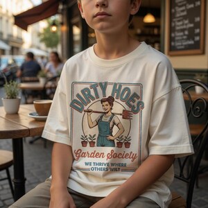 May include: Cream-coloured t-shirt with a retro graphic. The graphic features the text "Dirty Hoes Garden Society" with an illustration of a woman holding a garden tool. Below the graphic is the text "We thrive where others wilt."
