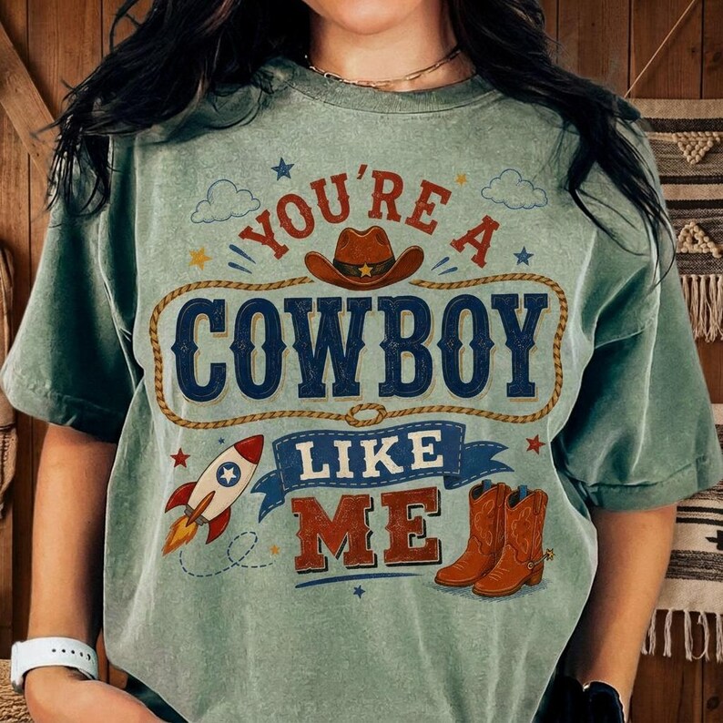 May include: A sage green short-sleeve t-shirt featuring a graphic print with the text "YOU'RE A COWBOY LIKE ME". The design includes a brown cowboy hat, a lasso, a rocket ship, and a pair of brown cowboy boots, accented with small blue stars.