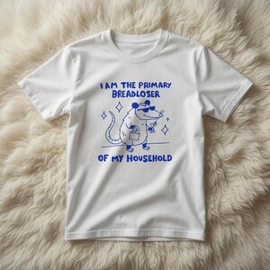 May include: White t-shirt with a blue graphic of a possum wearing sunglasses, holding a drink and a bag. The text above the possum reads "I AM THE PRIMARY BREADLOSER" and below it reads "OF MY HOUSEHOLD".