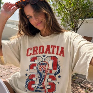 May include: An off-white short-sleeve t-shirt featuring a distressed red and blue graphic print. The text reads CROATIA above the year 2026, with a central illustration of a trophy containing a checkered emblem and small blue stars.