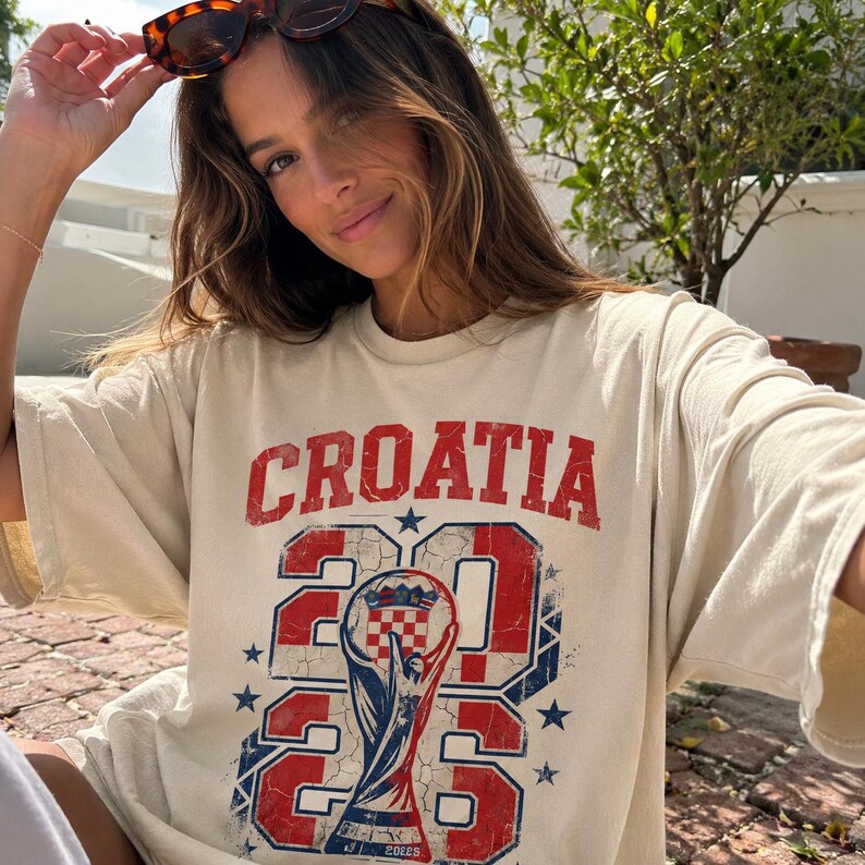 May include: An off-white short-sleeve t-shirt featuring a distressed red and blue graphic print. The text reads CROATIA above the year 2026, with a central illustration of a trophy containing a checkered emblem and small blue stars.