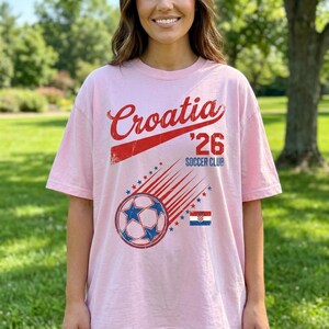 May include: A light pink short-sleeve t-shirt featuring a red graphic that reads Croatia '26 Soccer Club. The design includes a soccer ball with star patterns, motion lines, and a small rectangular Croatian flag in red, white, and blue colors.