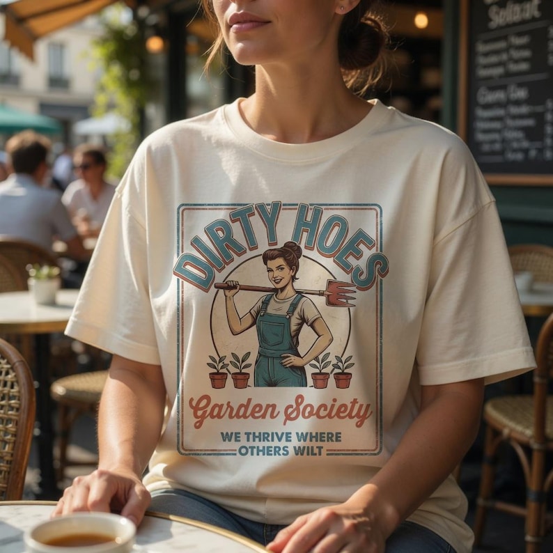 May include: Cream-colored t-shirt featuring a vintage-style graphic. The design includes the words "DIRTY HOES" in a circular frame, with a woman holding a gardening tool. Below, it reads "Garden Society" and "WE THRIVE WHERE OTHERS WILT".