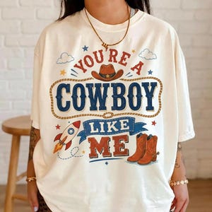 May include: An off-white short-sleeve t-shirt featuring a graphic print with the text "YOU'RE A COWBOY LIKE ME". The design includes a brown cowboy hat, a rocket ship, and a pair of brown cowboy boots, all surrounded by small stars and clouds.