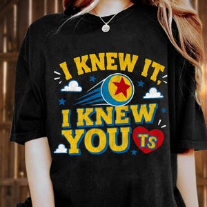 K&ouml;nnte beinhalten: Ein schwarzes Kurzarm-T-Shirt mit dem gelben Text I KNEW IT, I KNEW YOU und TS in einem roten Herz. Das Design enth&auml;lt einen blau-gelben Ball mit einem roten Stern, blaue Bewegungslinien sowie kleine wei&szlig;e Wolken und blaue Sterne.
