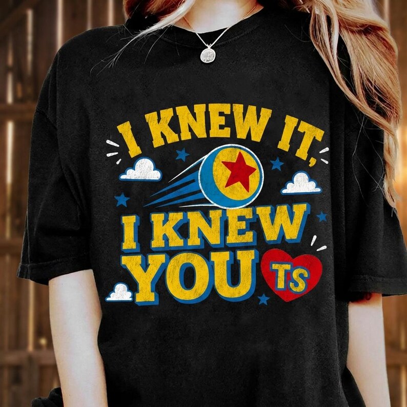 K&ouml;nnte beinhalten: Ein schwarzes Kurzarm-T-Shirt mit dem gelben Text I KNEW IT, I KNEW YOU und TS in einem roten Herz. Das Design enth&auml;lt einen blau-gelben Ball mit einem roten Stern, blaue Bewegungslinien sowie kleine wei&szlig;e Wolken und blaue Sterne.