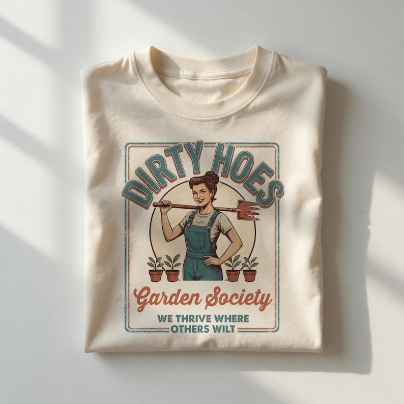 May include: A cream-coloured t-shirt with a vintage-style graphic. The design features a woman holding a gardening tool, the text "Dirty Hoes Garden Society", and the slogan "We thrive where others wilt".