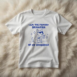 May include: White t-shirt with a blue graphic of a dog wearing sunglasses and holding a cocktail. The text on the shirt reads "I AM THE PRIMARY BREADLOSER OF MY HOUSEHOLD."