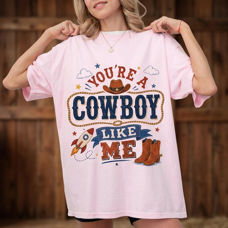 May include: A light pink t-shirt featuring a graphic print with the text "YOU'RE A COWBOY LIKE ME". The design includes a brown cowboy hat, a lasso, a rocket ship, and a pair of brown cowboy boots, accented with small stars and clouds.