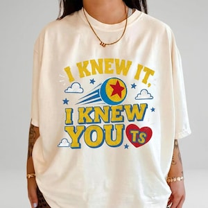 K&ouml;nnte beinhalten: Ein cremefarbenes T-Shirt mit kurzen &Auml;rmeln und dem gelben Schriftzug I KNEW IT, I KNEW YOU sowie einem roten Herz mit den Buchstaben TS. Das Design zeigt einen blauen Ball mit rotem Stern, blauen Bewegungslinien, kleinen Sternen und Wolken.