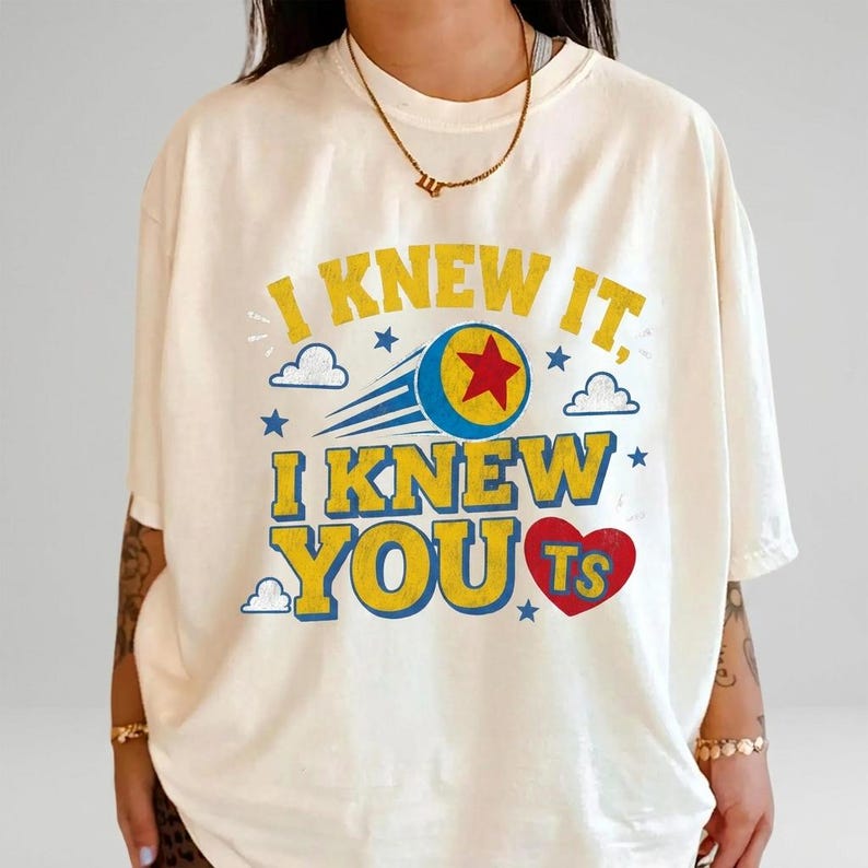 K&ouml;nnte beinhalten: Ein cremefarbenes T-Shirt mit kurzen &Auml;rmeln und dem gelben Schriftzug I KNEW IT, I KNEW YOU sowie einem roten Herz mit den Buchstaben TS. Das Design zeigt einen blauen Ball mit rotem Stern, blauen Bewegungslinien, kleinen Sternen und Wolken.