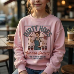 May include: Pale pink sweatshirt with the words "DIRTY HOES" in a retro design. The graphic shows a woman with a gardening tool, with "Garden Society" and "WE THRIVE WHERE OTHERS WILT" underneath. The sweatshirt is a light pink colour.