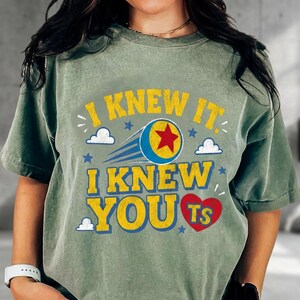 K&ouml;nnte beinhalten: Ein salbeigr&uuml;nes Kurzarm-T-Shirt mit dem gelben Text I KNEW IT, I KNEW YOU und TS in einem roten Herz. Das Design zeigt einen blauen und gelben Ball mit einem roten Stern, blauen Bewegungslinien sowie kleinen wei&szlig;en Wolken und blauen Sternen.