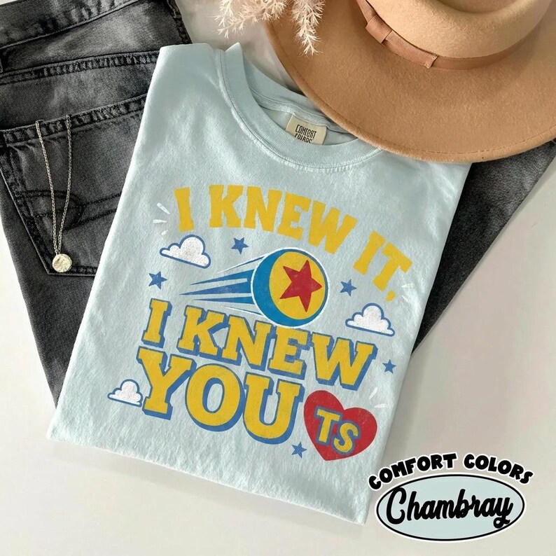 K&ouml;nnte beinhalten: Ein hellblaues T-Shirt mit dem gelben Text I KNEW IT, I KNEW YOU, einem roten Herz mit den Buchstaben TS, einem blau-gelben Ball mit einem roten Stern sowie kleinen wei&szlig;en Wolken und blauen Sternen. Das T-Shirt hat einen Rundhalsausschnitt.