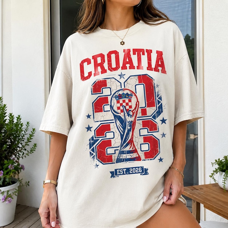 May include: An off-white short-sleeve t-shirt featuring a distressed red and blue graphic. The text reads CROATIA above the large numbers 2026, which frame a central trophy illustration containing a checkered pattern. Below is a banner with EST. 2026.