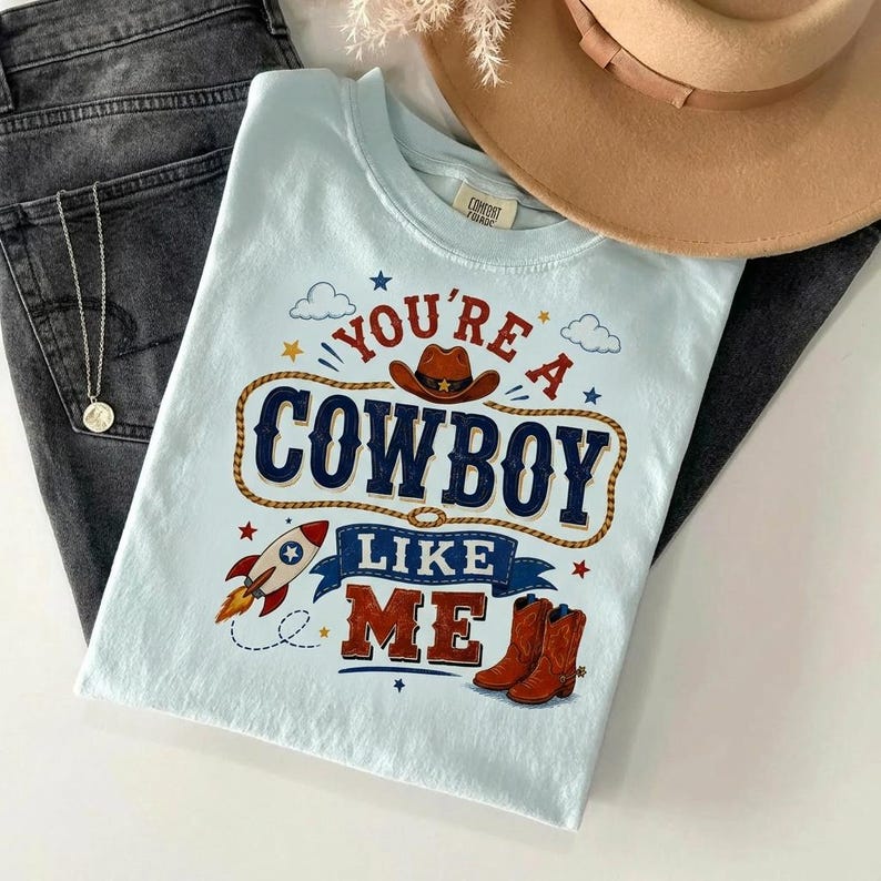 May include: A light blue t-shirt featuring the text "YOU'RE A COWBOY LIKE ME" in dark blue and red lettering. The graphic includes a brown cowboy hat, a rocket ship, and a pair of brown cowboy boots, all surrounded by small stars and clouds.