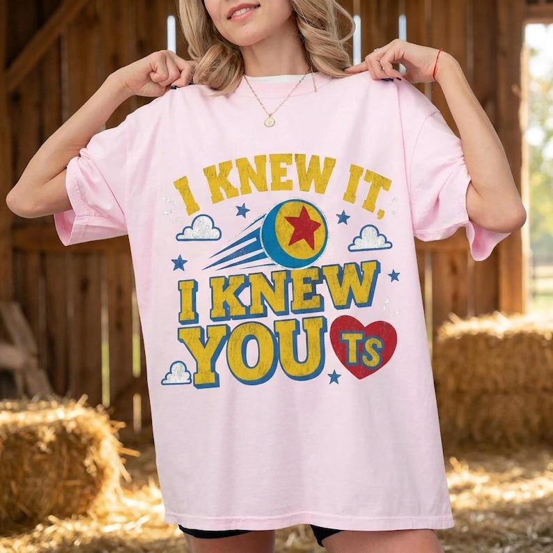 K&ouml;nnte beinhalten: Ein hellrosa T-Shirt mit kurzen &Auml;rmeln und einem Grafikdruck mit dem Text "I KNEW IT, I KNEW YOU" in gelben Buchstaben. Das Design enth&auml;lt einen blauen Ball mit einem roten Stern, kleine wei&szlig;e Wolken, blaue Sterne und ein rotes Herz mit den Buchstaben TS.