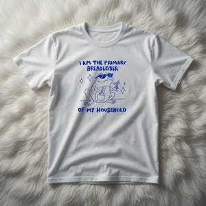 May include: White t-shirt with a blue graphic of a frog wearing sunglasses, holding a purse and a cocktail. The text above the frog reads "I AM THE PRIMARY BREADLOSER OF MY HOUSEHOLD."