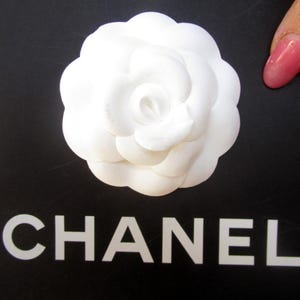 May include: A white camellia flower on a black surface with the word "CHANEL" in white block letters. The flower has multiple layers of petals. A pink fingernail is visible in the image.
